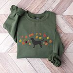 Military Green crewneck sweatshirt laid flat on herringbone wood, embroidered with a black Labrador silhouette under an arc of orange, red and gold autumn leaves across the chest