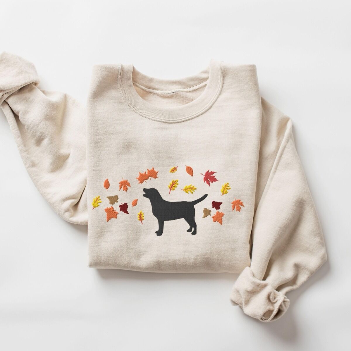 Black Lab Fall Sweatshirt – Sand Sand crewneck sweatshirt laid flat on white, embroidered with a black Labrador silhouette under an arc of orange, red and gold autumn leaves across the chest