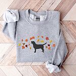 Sport Grey crewneck sweatshirt laid flat on herringbone wood, embroidered with a black Labrador silhouette under an arc of orange, red and gold autumn leaves across the chest