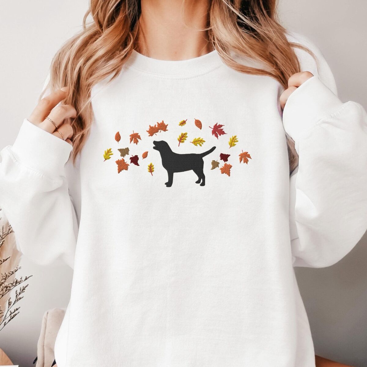 Black Lab Fall Sweatshirt - White Woman in a White crewneck sweatshirt embroidered with a black Labrador silhouette under an arc of orange, red and gold autumn leaves across the chest