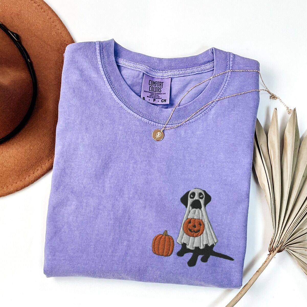Black Lab Ghost Shirt – Violet Black lab ghost shirt in Violet laid flat with a necklace and a felt hat, embroidered with a black Labrador in a white ghost sheet holding a jack-o'-lantern bucket beside a pumpkin