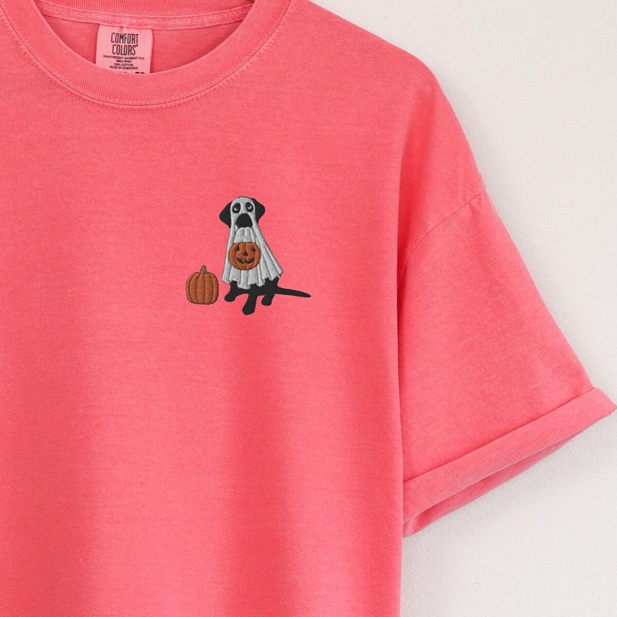 Black Lab Ghost Shirt - Watermelon Watermelon T-shirt on a wooden hanger with a rolled sleeve, embroidered with a black Labrador in a ghost sheet beside an orange pumpkin