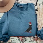 Indigo Blue crewneck sweatshirt laid flat beside a hat and pampas grass, embroidered with a small black Labrador in a Santa hat and red booties on the left chest