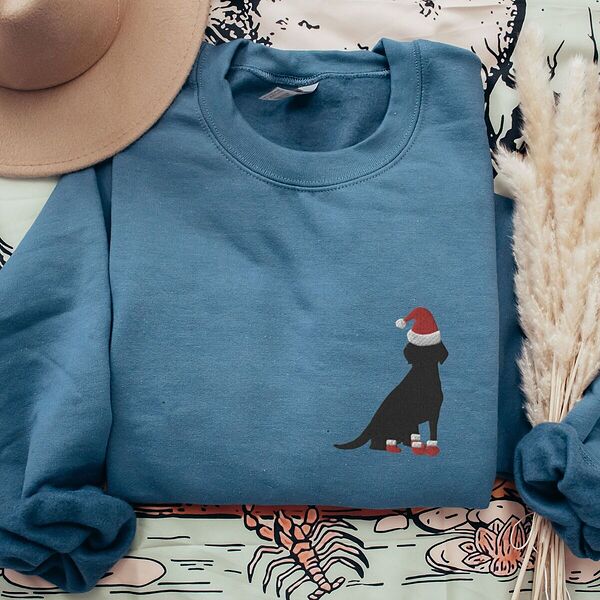 Indigo Blue crewneck sweatshirt laid flat beside a hat and pampas grass, embroidered with a small black Labrador in a Santa hat and red booties on the left chest