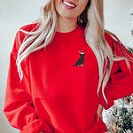 Woman by a Christmas tree in a Red crewneck sweatshirt embroidered with a small black Labrador in a Santa hat and red booties on the left chest