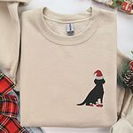 Sand crewneck sweatshirt folded with plaid ribbon and Christmas decorations, embroidered with a small black Labrador in a Santa hat and red booties on the left chest