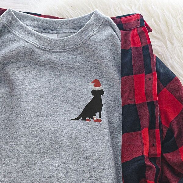 Sport Grey crewneck sweatshirt laid on a buffalo plaid shirt, embroidered with a small black Labrador in a Santa hat and red booties on the left chest