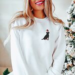 Woman smiling by a Christmas tree in a White crewneck sweatshirt embroidered with a small black Labrador in a Santa hat and red booties on the left chest