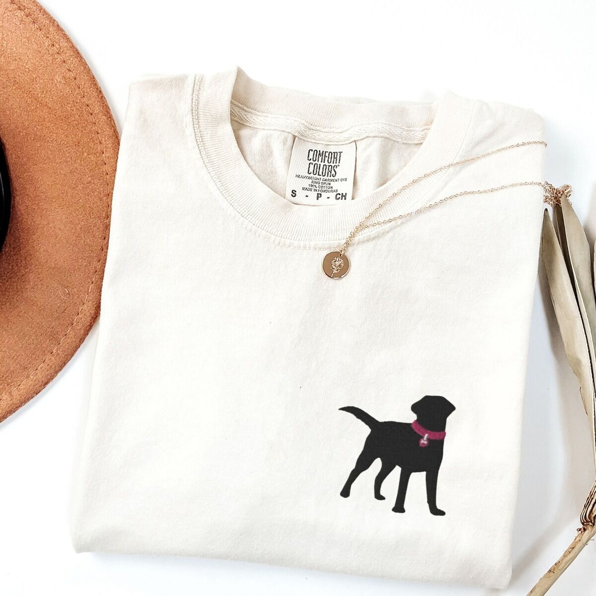 Black Lab Silhouette Shirt – Ivory Ivory T-shirt laid flat with a hat and a necklace, embroidered with a small black Labrador silhouette in a hot pink collar and tag on the left chest