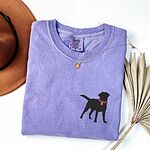 Violet T-shirt laid flat with a hat and a gold necklace, embroidered with a small black Labrador silhouette in a hot pink collar and tag on the left chest