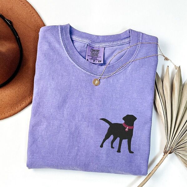 Violet T-shirt laid flat with a hat and a gold necklace, embroidered with a small black Labrador silhouette in a hot pink collar and tag on the left chest