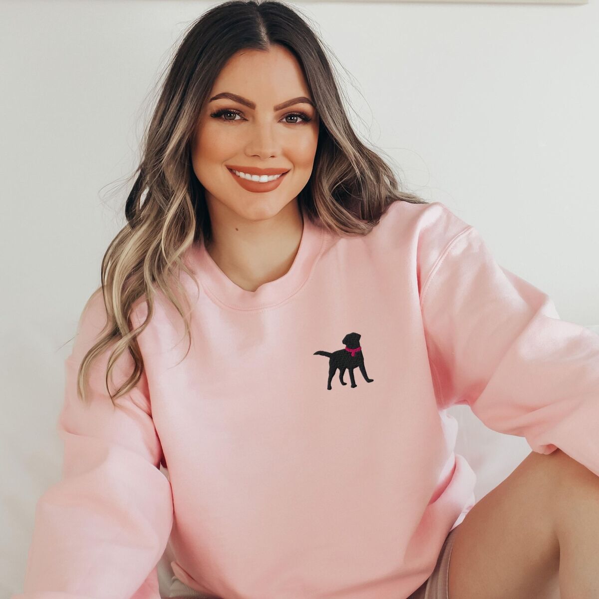 Black Lab Silhouette Sweatshirt - Light Pink Smiling woman in a Light Pink crewneck sweatshirt embroidered with a small black Labrador silhouette in a pink collar on the left chest