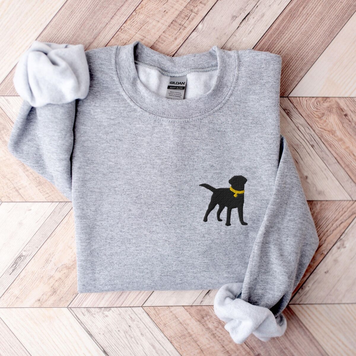 Black Lab Silhouette Sweatshirt - Sport Grey Sport Grey crewneck sweatshirt laid flat on herringbone wood, embroidered with a small black Labrador silhouette in a yellow collar