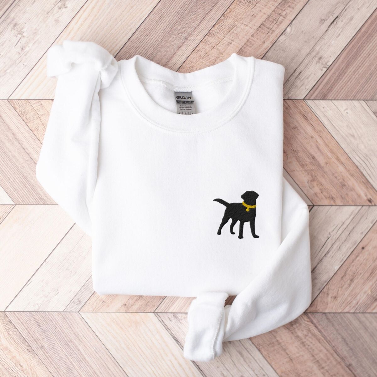 Black Lab Silhouette Sweatshirt - White White crewneck sweatshirt laid flat on herringbone wood, embroidered with a small black Labrador silhouette in a yellow collar on the left chest