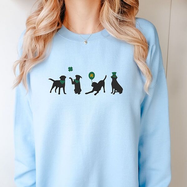 Smiling woman in a Light Blue crewneck sweatshirt embroidered with four black lab silhouettes in green bandanas with a shamrock, a clover balloon and a leprechaun hat
