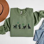 Military Green crewneck sweatshirt laid flat with a felt hat and jeans, embroidered with four black lab silhouettes in green bandanas with a shamrock, a clover balloon and a leprechaun hat
