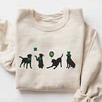 Sand crewneck sweatshirt laid flat, embroidered with four black lab silhouettes in green bandanas with a shamrock, a clover balloon and a leprechaun hat