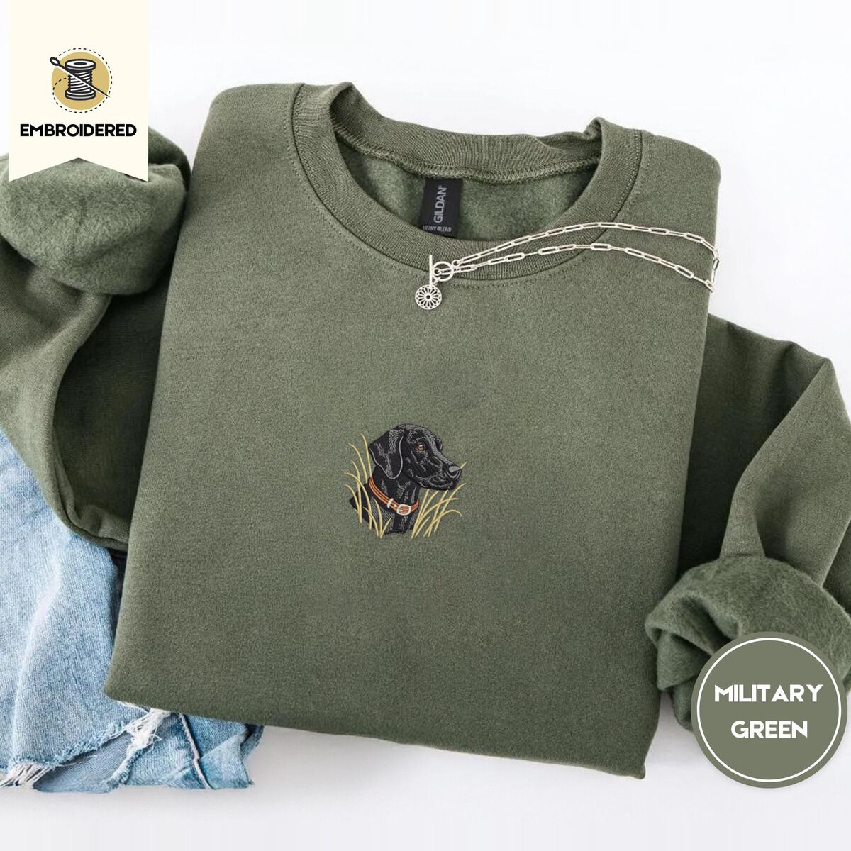 Black Lab Sweatshirt – Military Green Military Green crewneck sweatshirt folded with light jeans and a silver chain necklace, embroidered with a black Labrador head with a rust leather collar in tall marsh grass