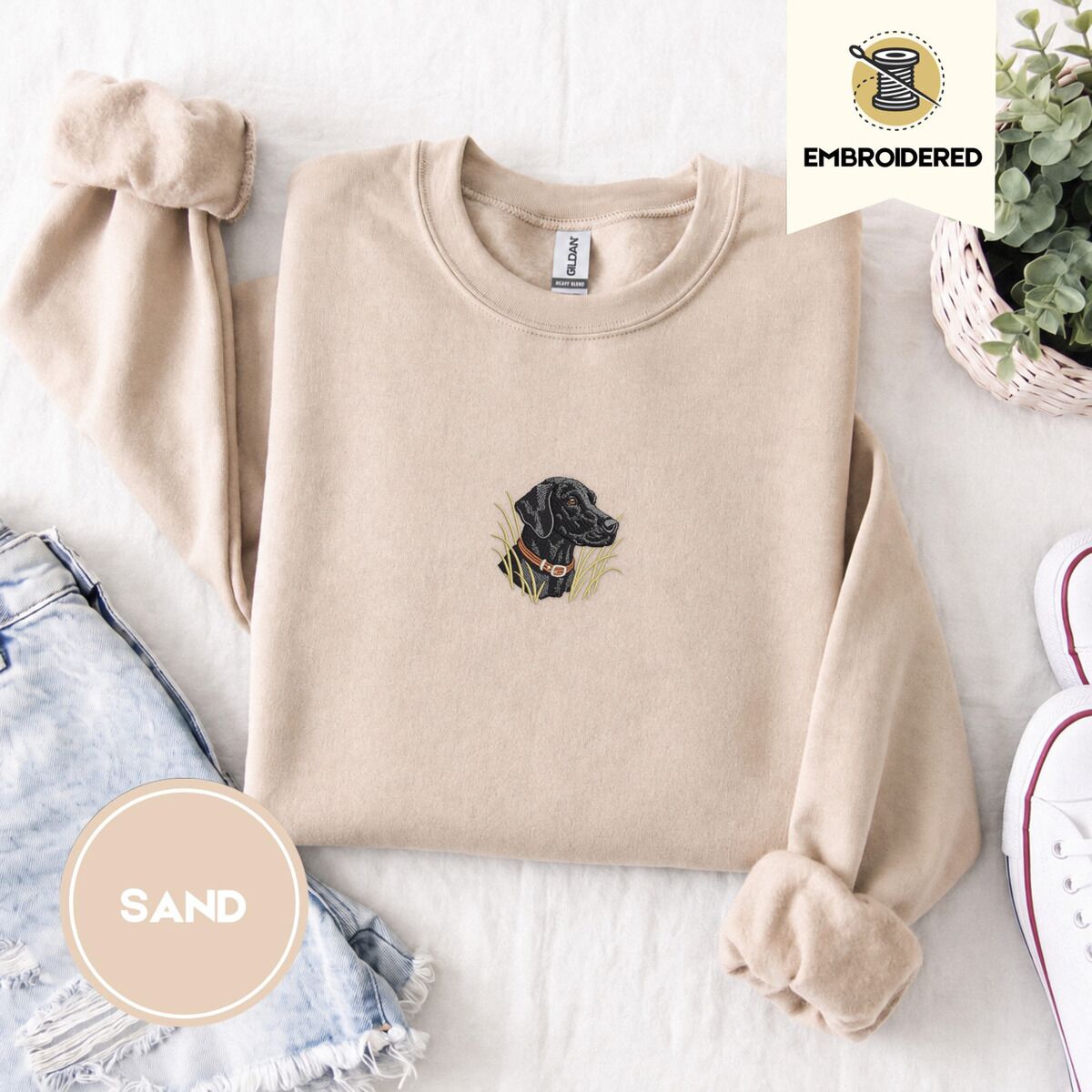 Black Lab Sweatshirt – Sand Sand crewneck sweatshirt folded with denim shorts, white sneakers and a potted plant, embroidered with a black Labrador head with a rust leather collar in tall marsh grass