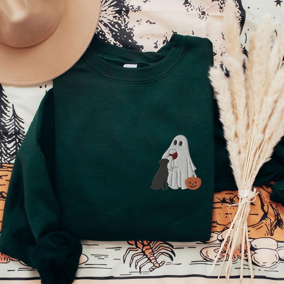 Black Lab Wine Sweatshirt – Forest Green Forest Green crewneck sweatshirt laid flat with a brown hat and pampas grass, embroidered with a small ghost lab mom sipping red wine with a black lab sitting against her and a jack-o'-lantern, on the left chest