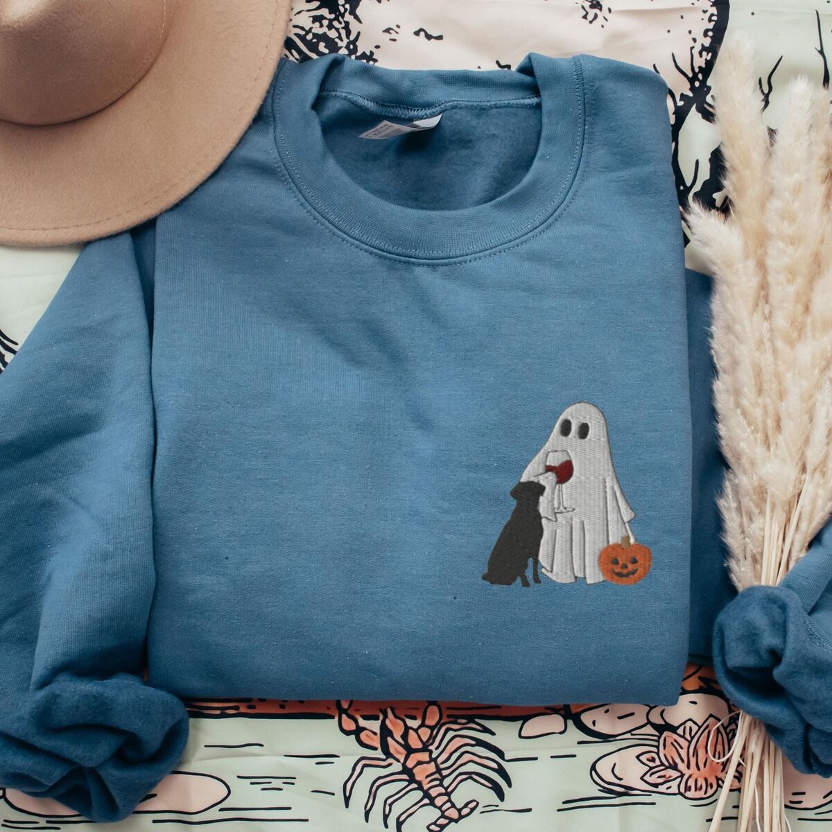 Black Lab Wine Sweatshirt – Indigo Blue Indigo Blue crewneck sweatshirt laid flat with a brown hat and pampas grass, embroidered with a small ghost lab mom sipping red wine with a black lab sitting against her and a jack-o'-lantern, on the left chest