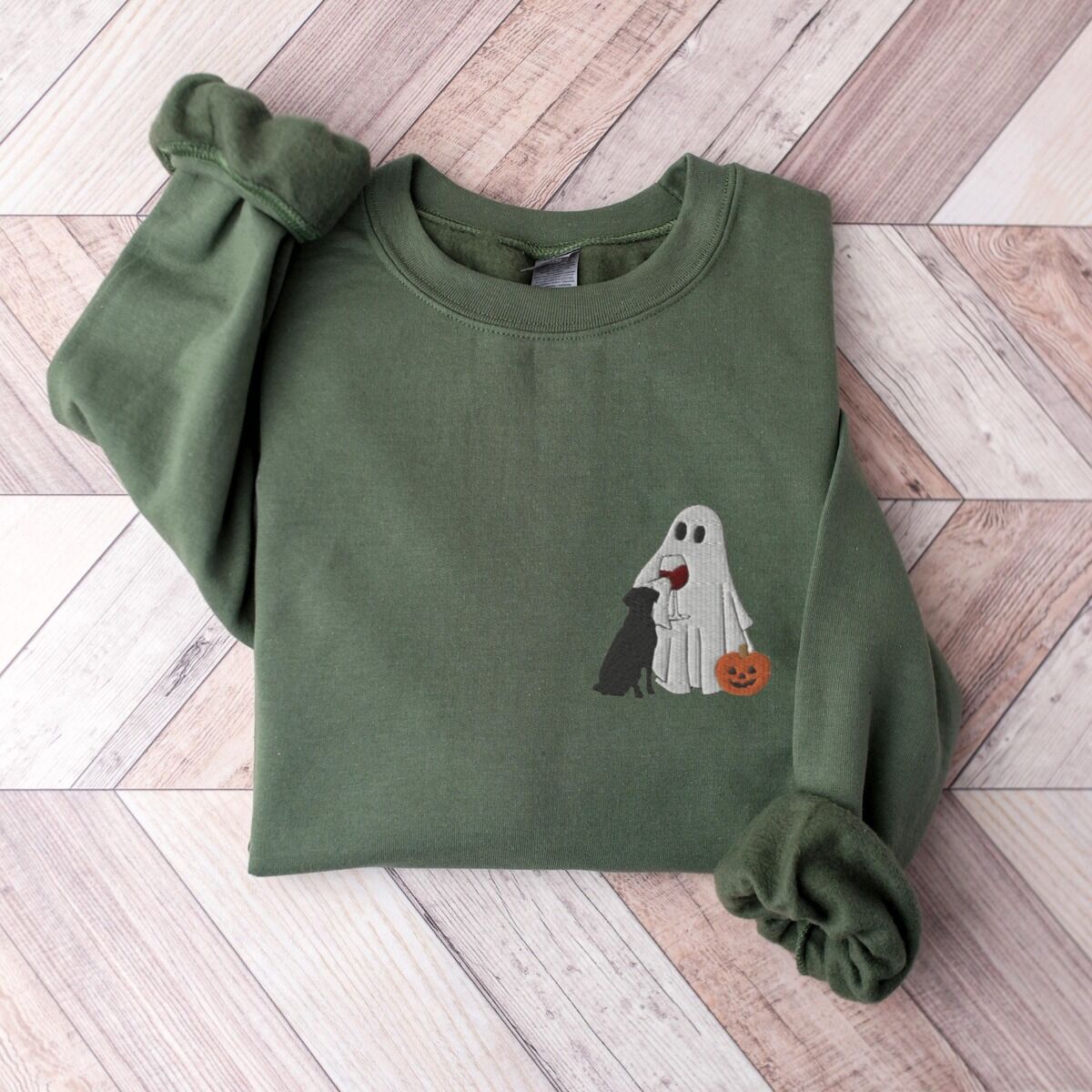Black Lab Wine Sweatshirt – Military Green Military Green crewneck sweatshirt laid flat on a wood floor, embroidered with a small ghost lab mom sipping red wine with a black lab sitting against her and a jack-o'-lantern, on the left chest