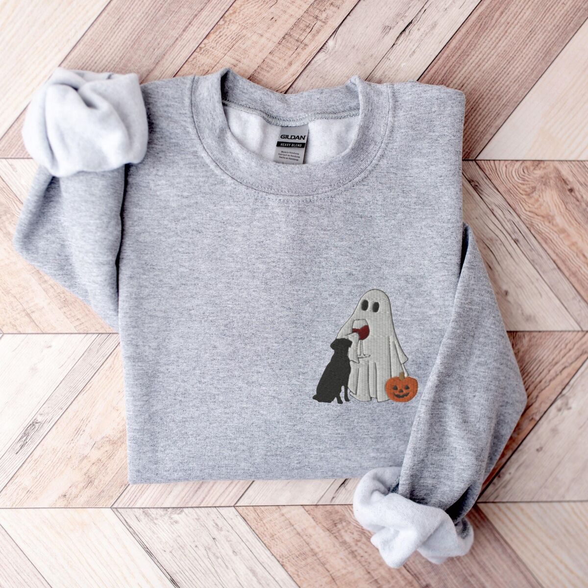 Black Lab Wine Sweatshirt - Sport Grey Sport Grey crewneck sweatshirt laid flat on a wood floor, embroidered with a small ghost lab mom sipping red wine with a black lab sitting against her and a jack-o'-lantern, on the left chest