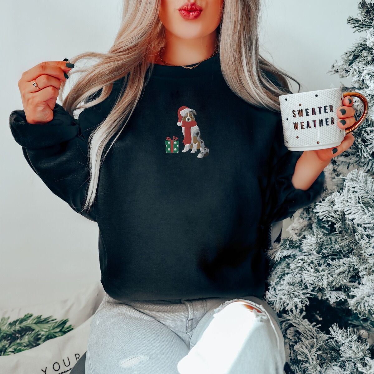 Blue Merle Aussie Christmas Sweatshirt – Black Woman holding a mug by a frosted Christmas tree in a Black crewneck sweatshirt embroidered with a blue merle Australian shepherd in a Santa hat and red scarf beside a green polka dot gift box