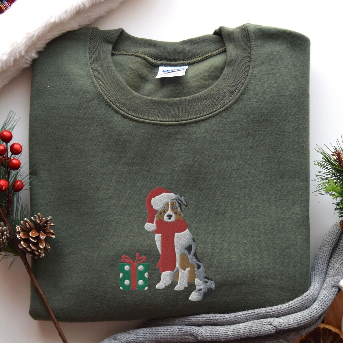 Blue Merle Aussie Christmas Sweatshirt – Military Green Military Green crewneck sweatshirt laid flat with pine cones, red berries and a grey knit blanket, embroidered with a blue merle Australian shepherd in a Santa hat and red scarf beside a green polka dot gift box