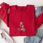 Red crewneck sweatshirt laid flat with jeans and white sneakers, embroidered with a blue merle Australian shepherd in a Santa hat and red scarf beside a green polka dot gift box
