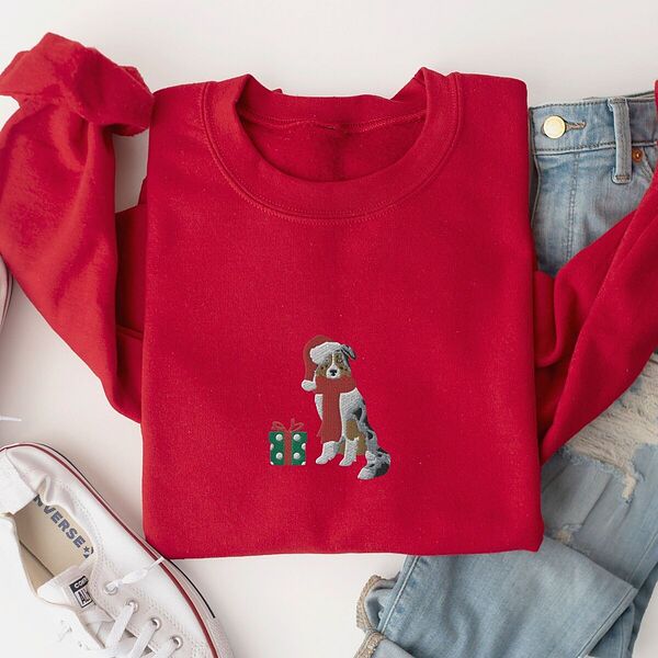 Red crewneck sweatshirt laid flat with jeans and white sneakers, embroidered with a blue merle Australian shepherd in a Santa hat and red scarf beside a green polka dot gift box