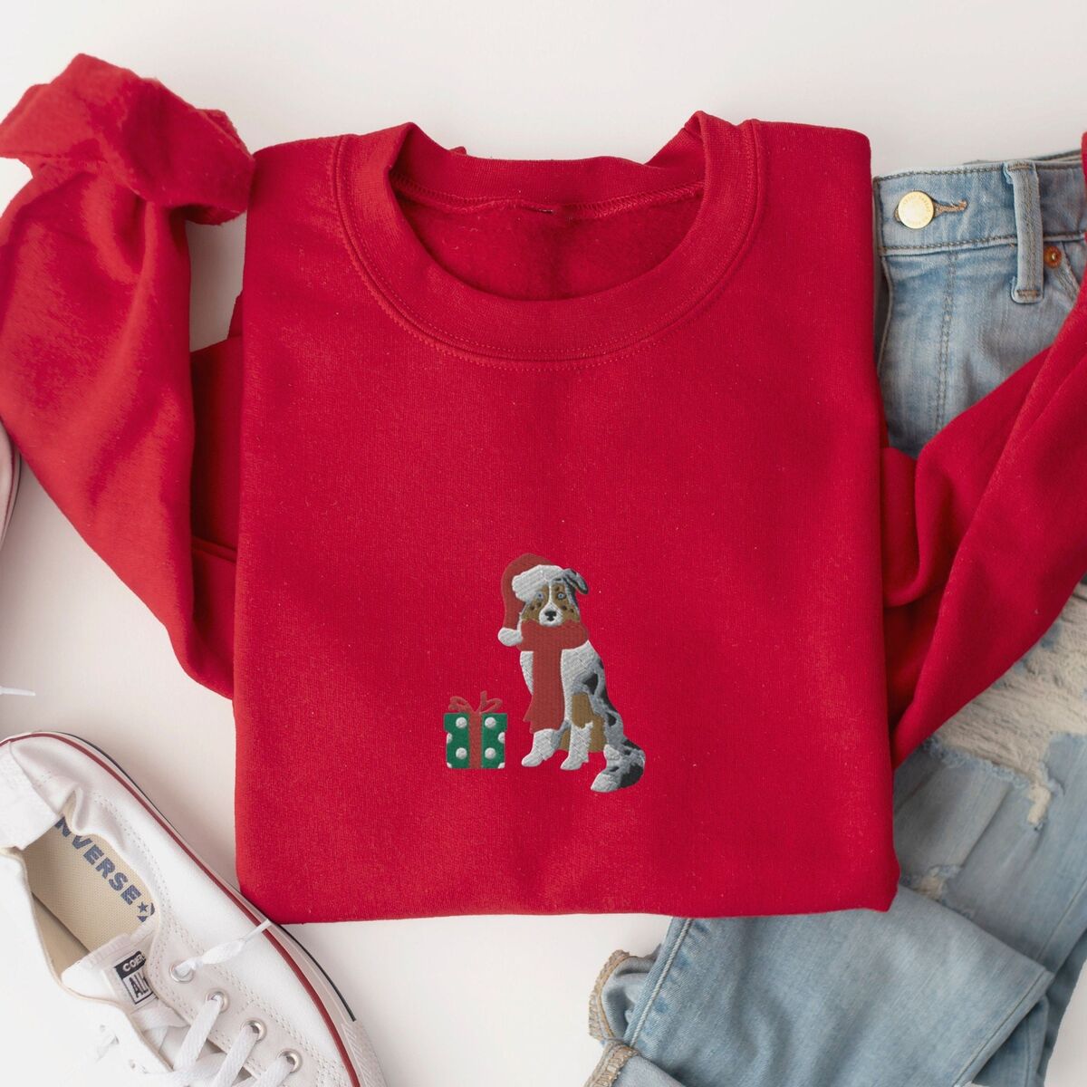 Blue Merle Aussie Christmas Sweatshirt - Red Red crewneck sweatshirt laid flat with jeans and white sneakers, embroidered with a blue merle Australian shepherd in a Santa hat and red scarf beside a green polka dot gift box