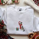 White crewneck sweatshirt laid flat with pine cones, red berries and dried orange slices, embroidered with a blue merle Australian shepherd in a Santa hat and red scarf beside a green polka dot gift box
