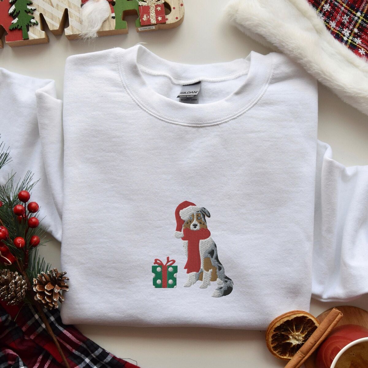 Blue Merle Aussie Christmas Sweatshirt - White White crewneck sweatshirt laid flat with pine cones, red berries and dried orange slices, embroidered with a blue merle Australian shepherd in a Santa hat and red scarf beside a green polka dot gift box