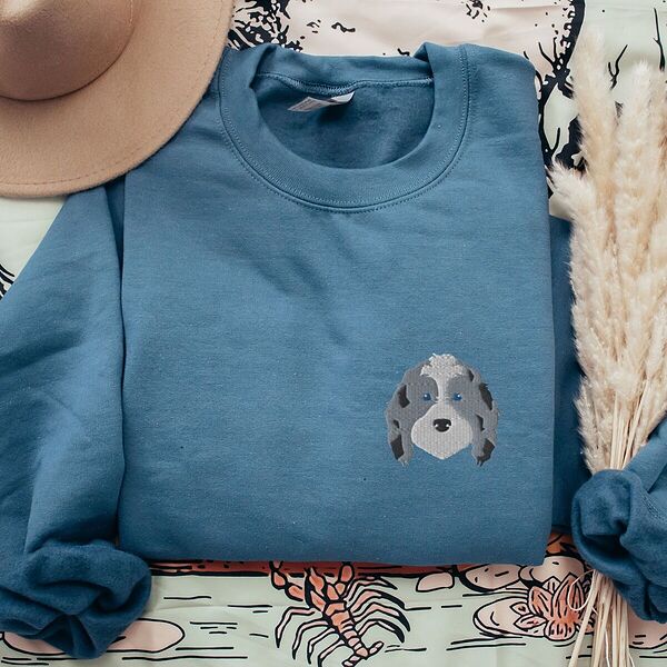 Indigo Blue crewneck sweatshirt laid flat with a tan hat and dried pampas grass, embroidered with a blue merle aussiedoodle face with a white blaze, blue eyes and spotted ears on the left chest