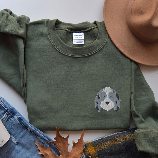 Military Green crewneck sweatshirt laid flat with a tan hat, jeans, a plaid scarf and autumn leaves, embroidered with a blue merle aussiedoodle face with a white blaze, blue eyes and spotted ears on the left chest
