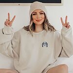 Woman in a beanie making peace signs in a Sand crewneck sweatshirt embroidered with a blue merle aussiedoodle face with a white blaze, blue eyes and spotted ears on the left chest