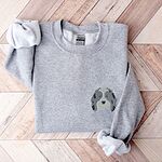 Sport Grey crewneck sweatshirt laid flat on a herringbone wood floor, embroidered with a blue merle aussiedoodle face with a white blaze, blue eyes and spotted ears on the left chest