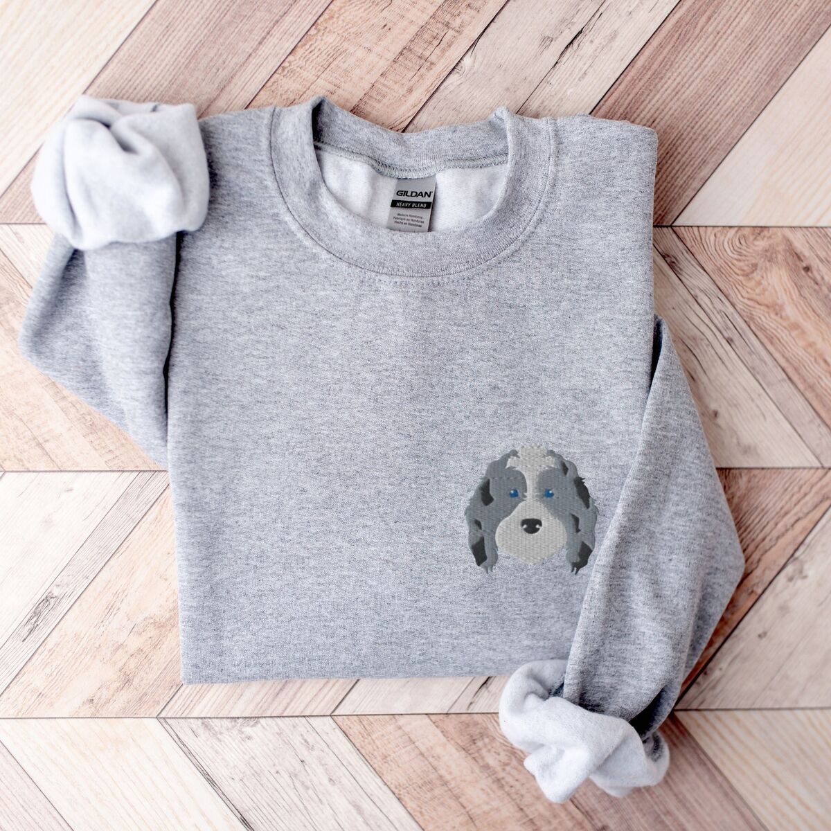 Blue Merle Aussiedoodle Sweatshirt - Sport Grey Sport Grey crewneck sweatshirt laid flat on a herringbone wood floor, embroidered with a blue merle aussiedoodle face with a white blaze, blue eyes and spotted ears on the left chest