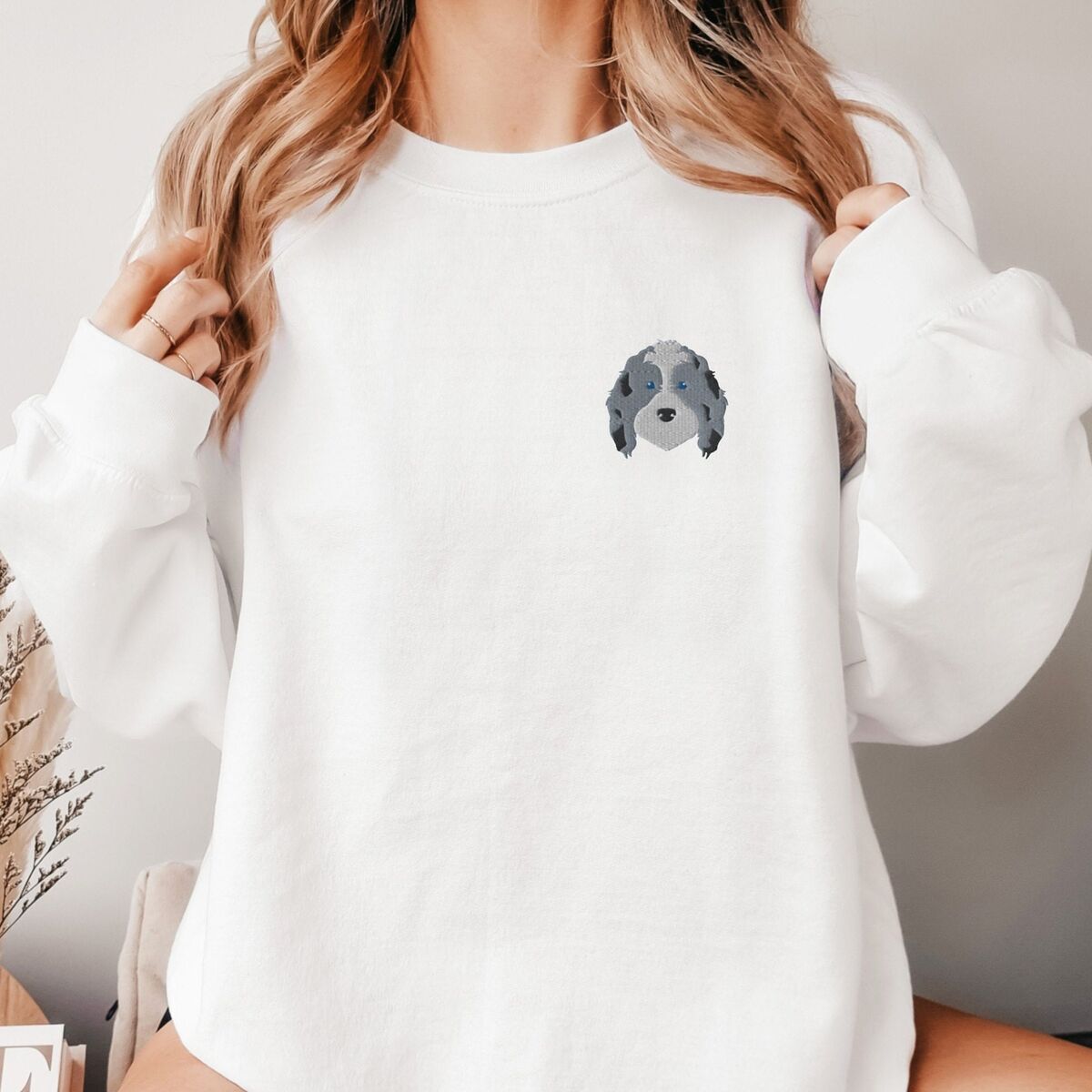 Blue Merle Aussiedoodle Sweatshirt - White Woman holding her hair in a White crewneck sweatshirt embroidered with a blue merle aussiedoodle face with a white blaze, blue eyes and spotted ears on the left chest