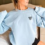 Woman sitting in a light blue crewneck sweatshirt embroidered with a small minimalist blue-grey pit bull face with a pale grey blaze on the left chest