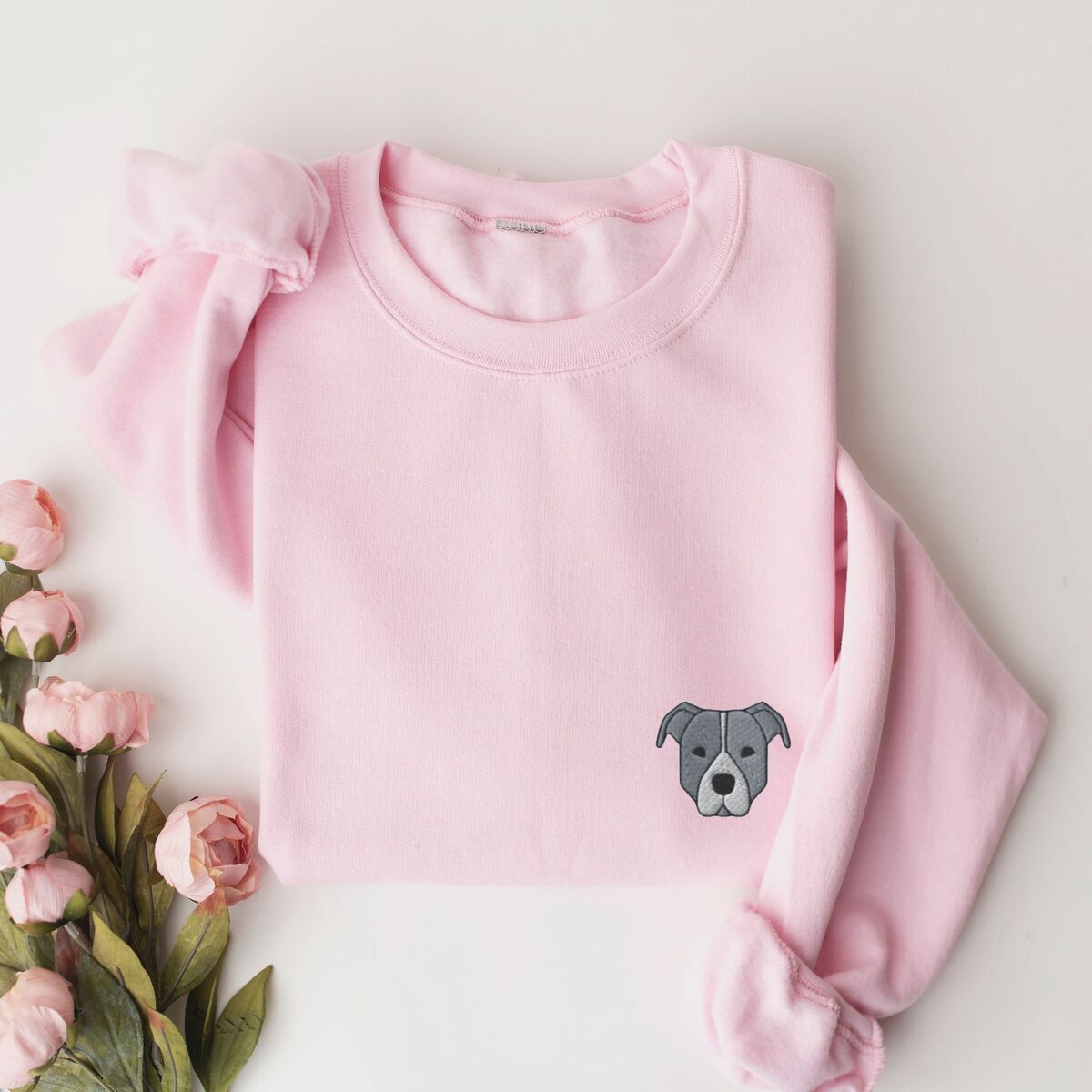 Blue Pitbull Face Sweatshirt - Light Pink Light Pink crewneck sweatshirt laid flat with pink roses, embroidered with a small minimalist blue-grey pit bull face with a pale grey blaze on the left chest