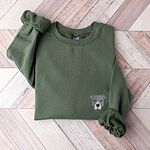 Military Green crewneck sweatshirt laid flat on a wood floor, embroidered with a small minimalist blue-grey pit bull face with a pale grey blaze on the left chest