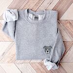 Sport Grey crewneck sweatshirt laid flat on a wood floor, embroidered with a small minimalist blue-grey pit bull face with a pale grey blaze on the left chest