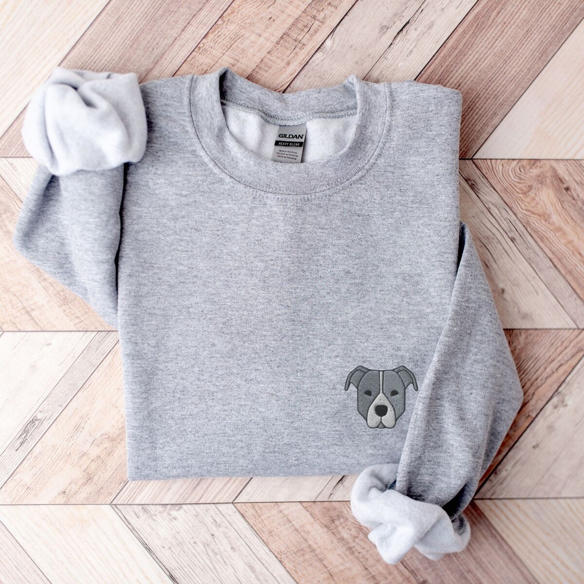 Blue Pitbull Face Sweatshirt - Sport Grey Sport Grey crewneck sweatshirt laid flat on a wood floor, embroidered with a small minimalist blue-grey pit bull face with a pale grey blaze on the left chest