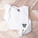 White crewneck sweatshirt laid flat on a wood floor, embroidered with a small minimalist blue-grey pit bull face with a pale grey blaze on the left chest