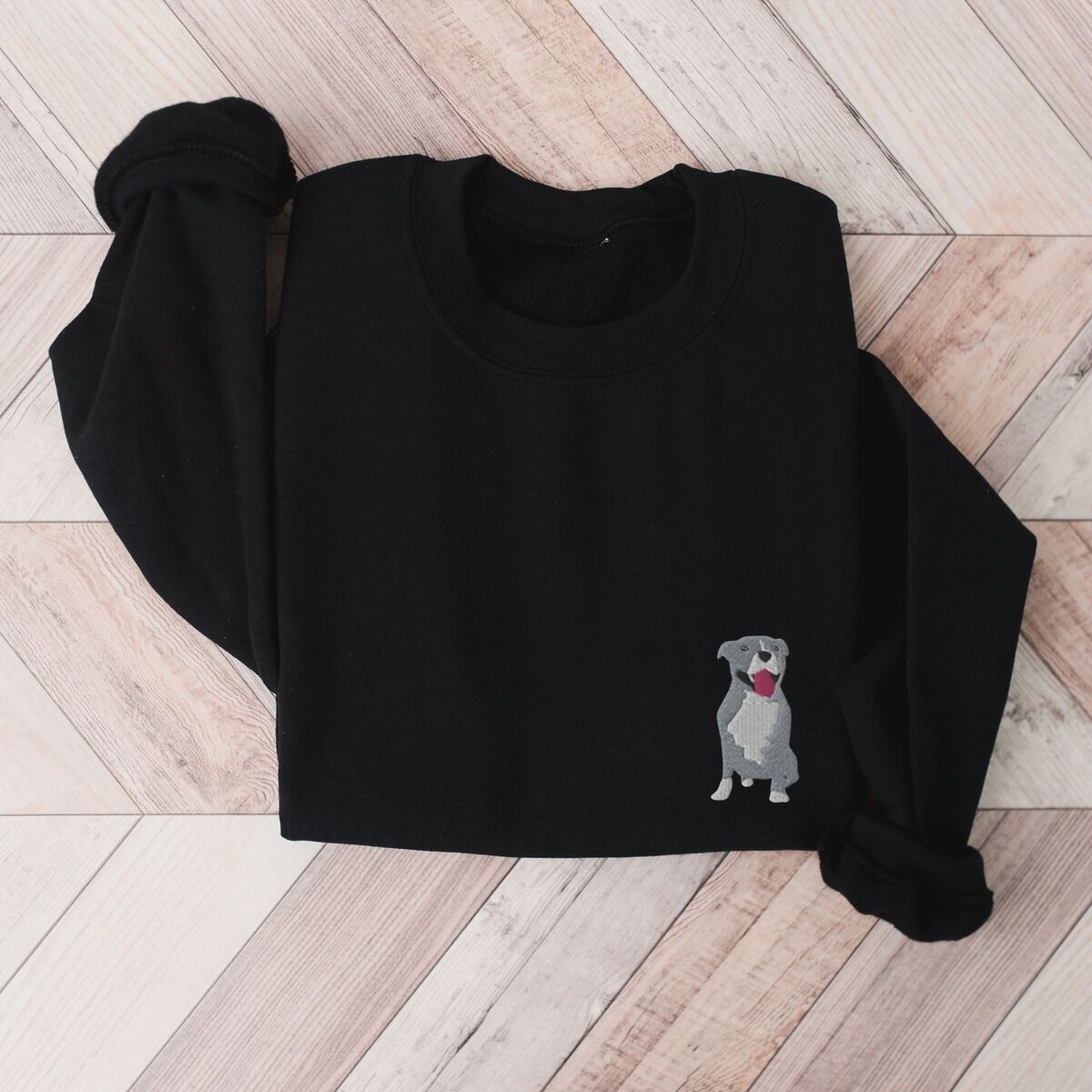 Blue Pitbull Sweatshirt – Black Black crewneck sweatshirt laid flat on a wood floor, embroidered with a small blue-grey pit bull sitting with its pink tongue out on the left chest