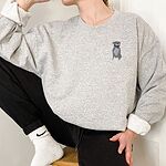 Woman sitting in a light grey crewneck sweatshirt, embroidered with a small blue-grey pit bull sitting with its pink tongue out on the left chest
