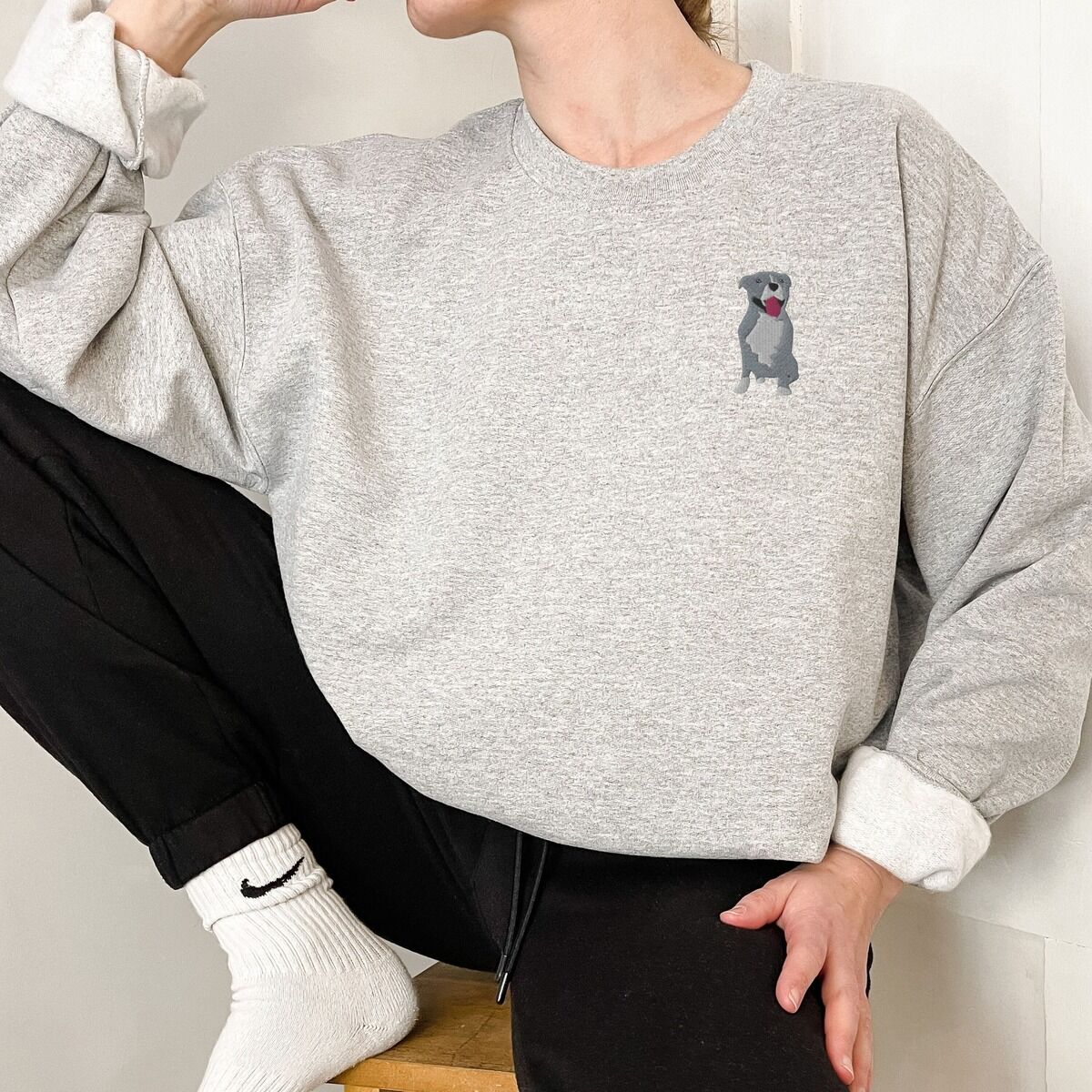 Blue Pitbull Sweatshirt - worn in grey Woman sitting in a light grey crewneck sweatshirt, embroidered with a small blue-grey pit bull sitting with its pink tongue out on the left chest