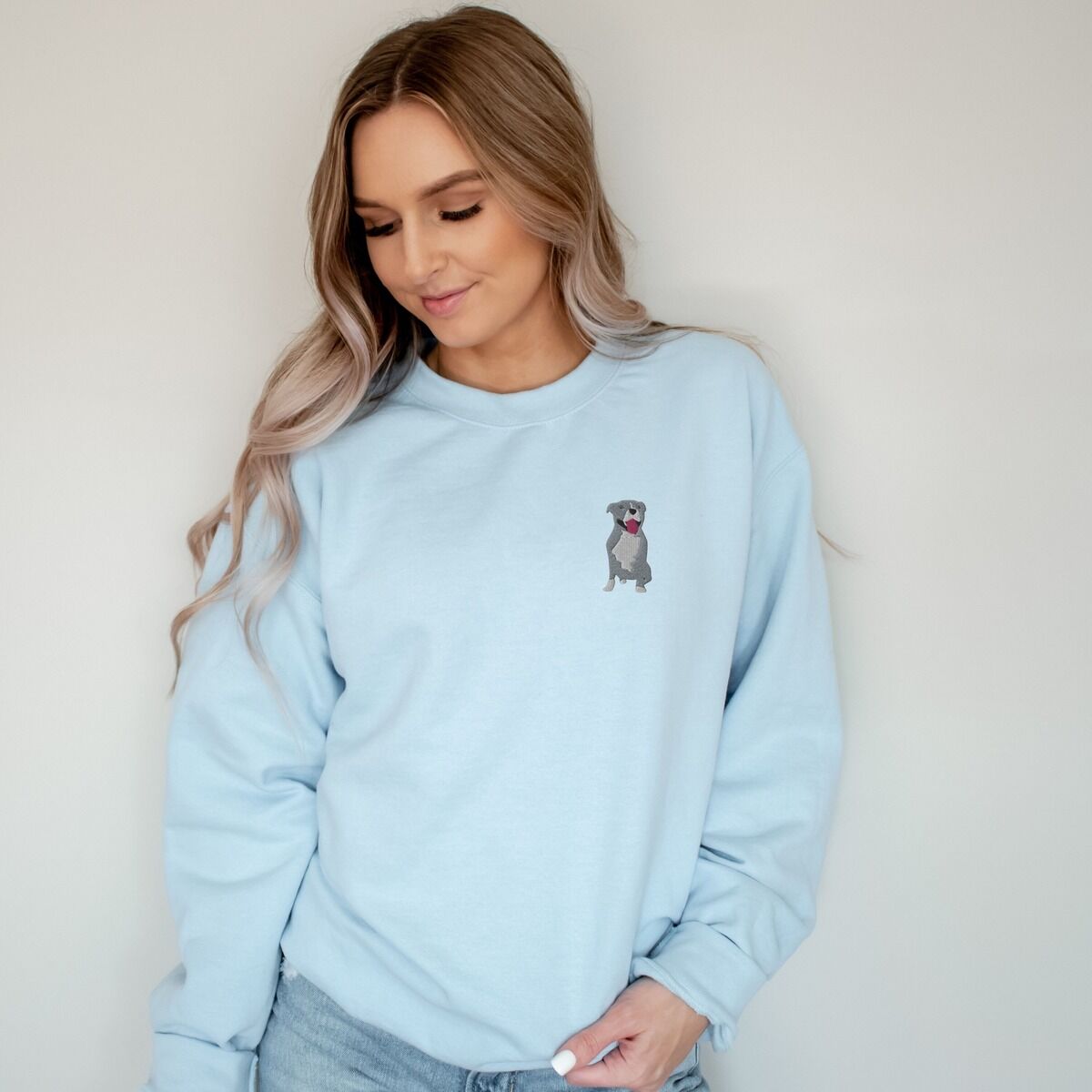 Blue Pitbull Sweatshirt - worn in light blue Woman in a light blue crewneck sweatshirt, embroidered with a small blue-grey pit bull sitting with its pink tongue out on the left chest